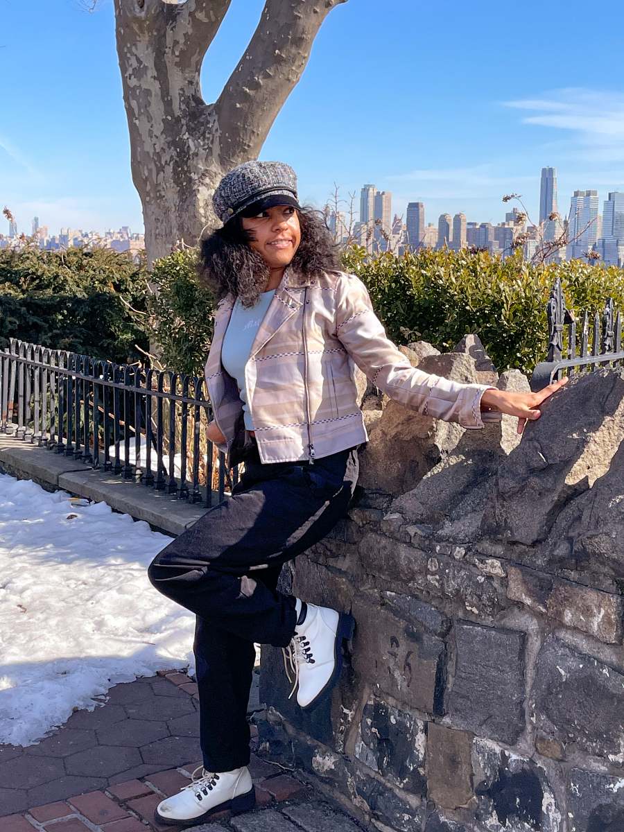 Woman holding a rock fence, leaning on the fence with her left leg bend, while looking sideways wearing baby-blue high neck baby tank top under a beige jacket with baggy black jeans and black-and-white boots.