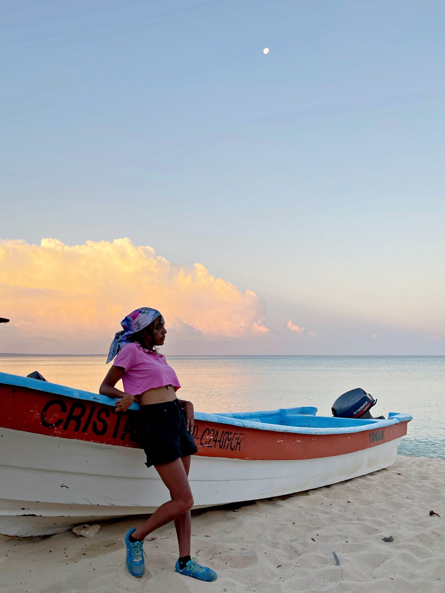Sunrise with the Caribbean ocean as a background. Girl leaning against a fishing boat wearing black shorts and a pink crop top 