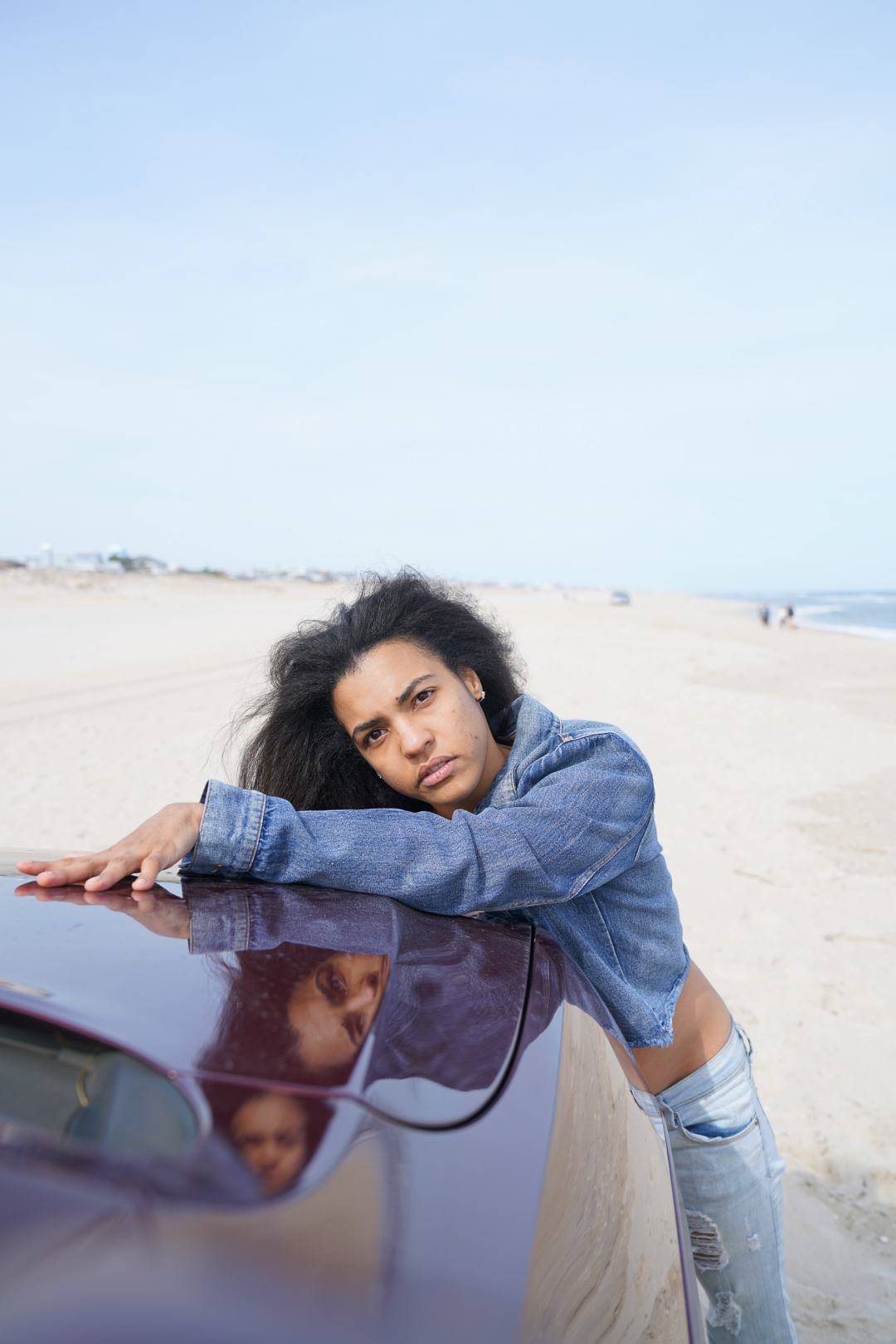 Brunette leaning upper body on the hood of the car looking straight at the camera with a serious face. She is wearing ripped light wash jeans and a jean jacket with her hair straight